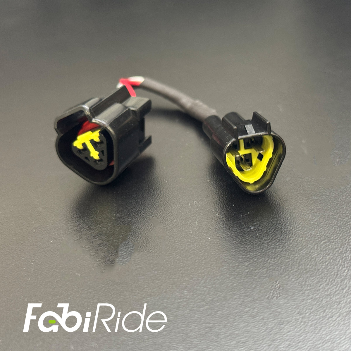 Elektrinio motociklo laidų adapteris su dviem juodomis jungtimis ir geltonais vidiniais kontaktais „FabiRide“ fone.
